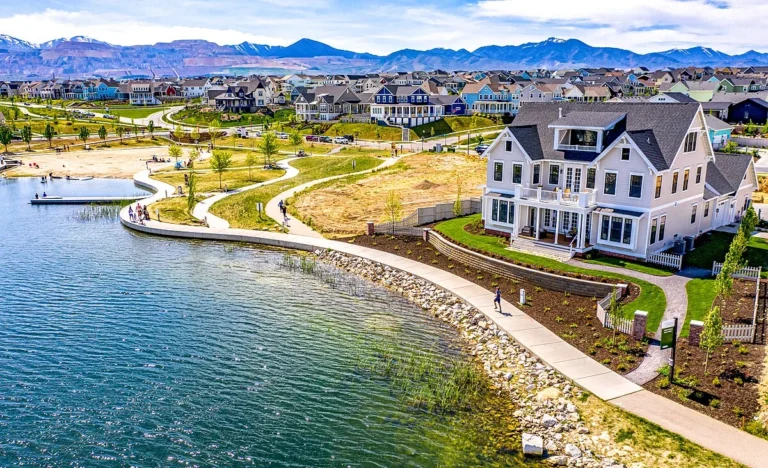 A scenic waterfront view in Daybreak, Utah, featuring a walking path, a large lake, modern homes, and mountain views in the background.