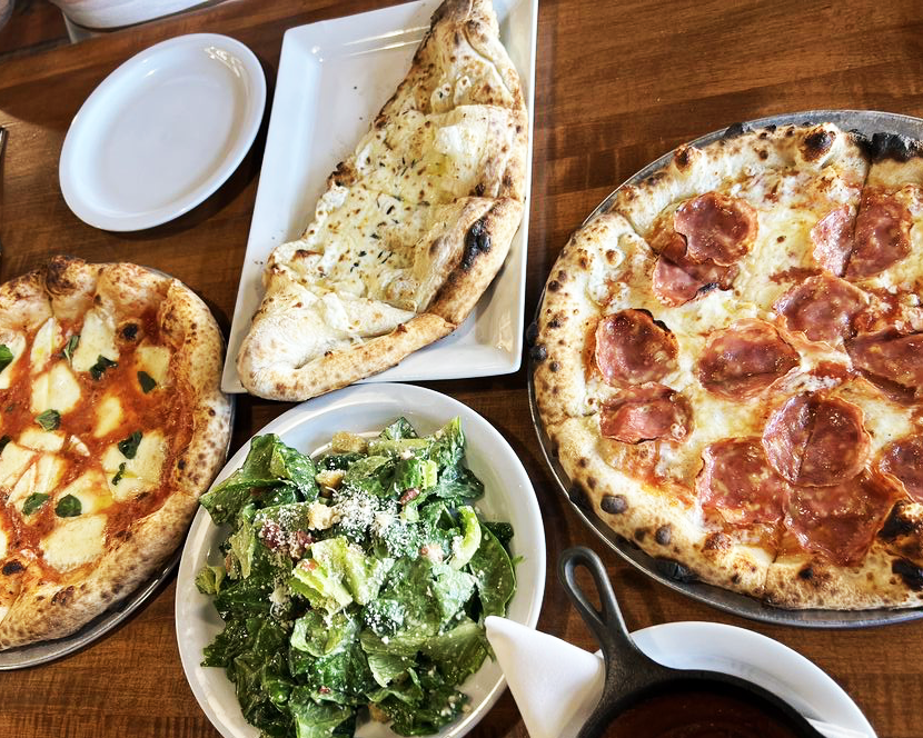 A table spread at Mozz Artisan Pizza in Daybreak, Utah, featuring a Margherita pizza, a pepperoni pizza, a cheesy calzone, and a fresh Caesar salad.