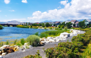 A scenic view of Oquirrh Lake in the middle of Daybreak, Utah, featuring walking trails, lush greenery, and homes lining the waterfront with mountains in the background.