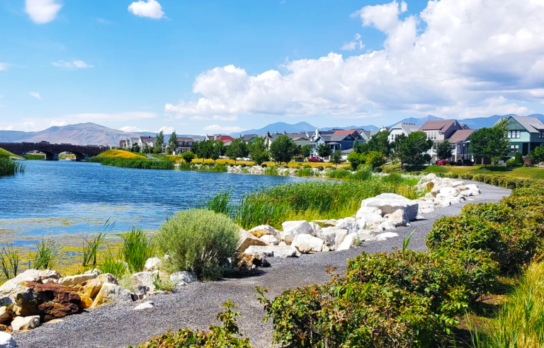 A scenic view of Oquirrh Lake in the middle of Daybreak, Utah, featuring walking trails, lush greenery, and homes lining the waterfront with mountains in the background.